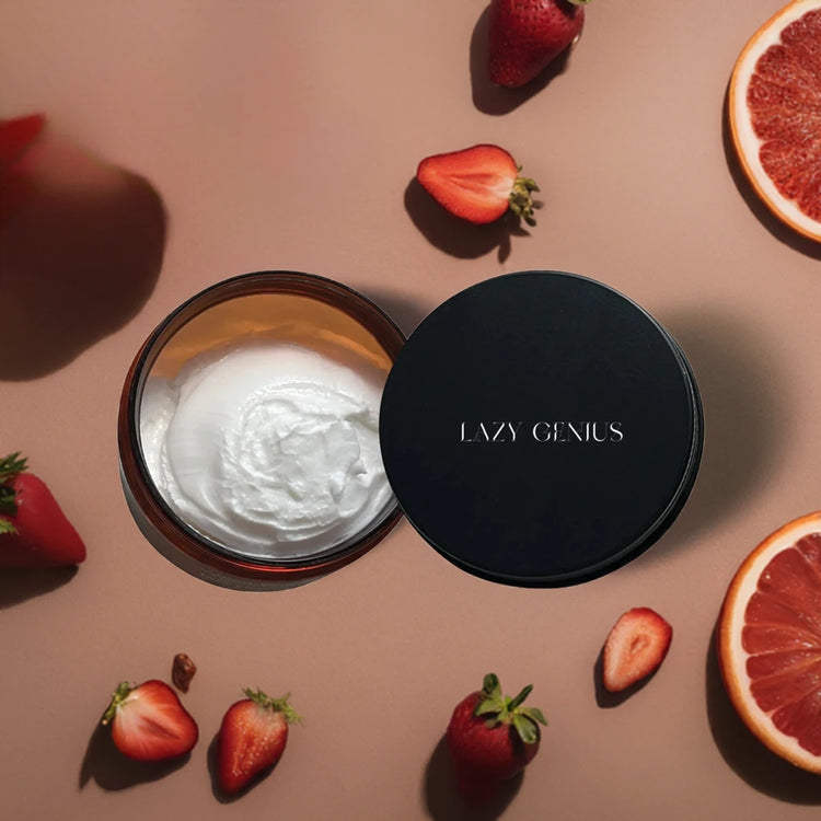 Open jar of Lazy Genius cream surrounded by fresh strawberries and grapefruit slices on a brown background.
